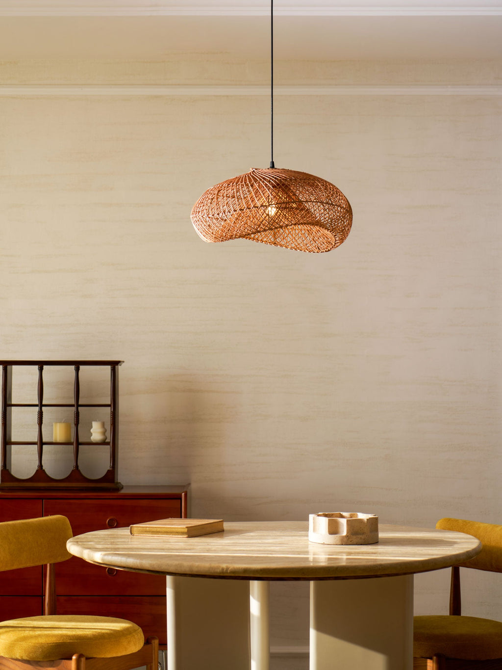 A Clérachic rattan pendant in the lower half of a cream panelled room, above a round table, with a red cabinet behind.