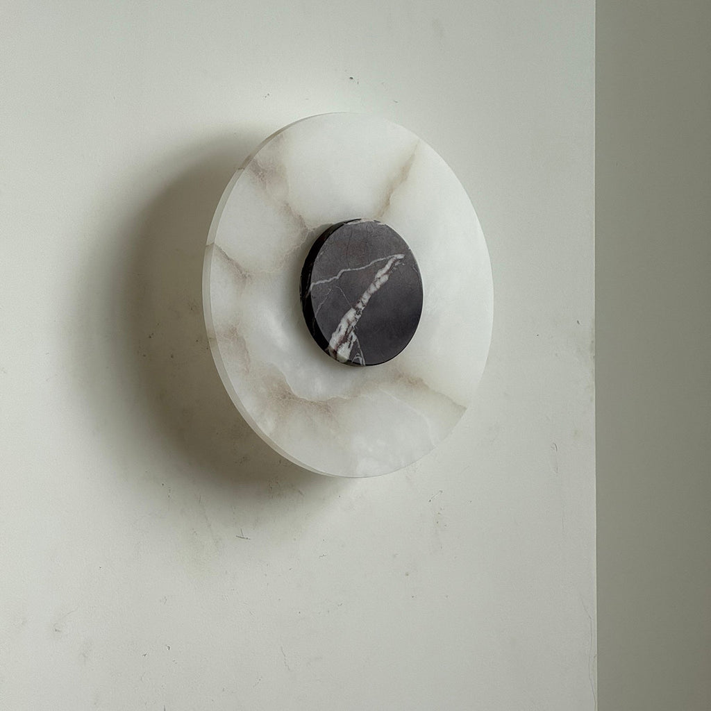 Clérachic wall lamp unlit on a pale wall, amber veining through the alabaster and a dark grey marble centre disc.