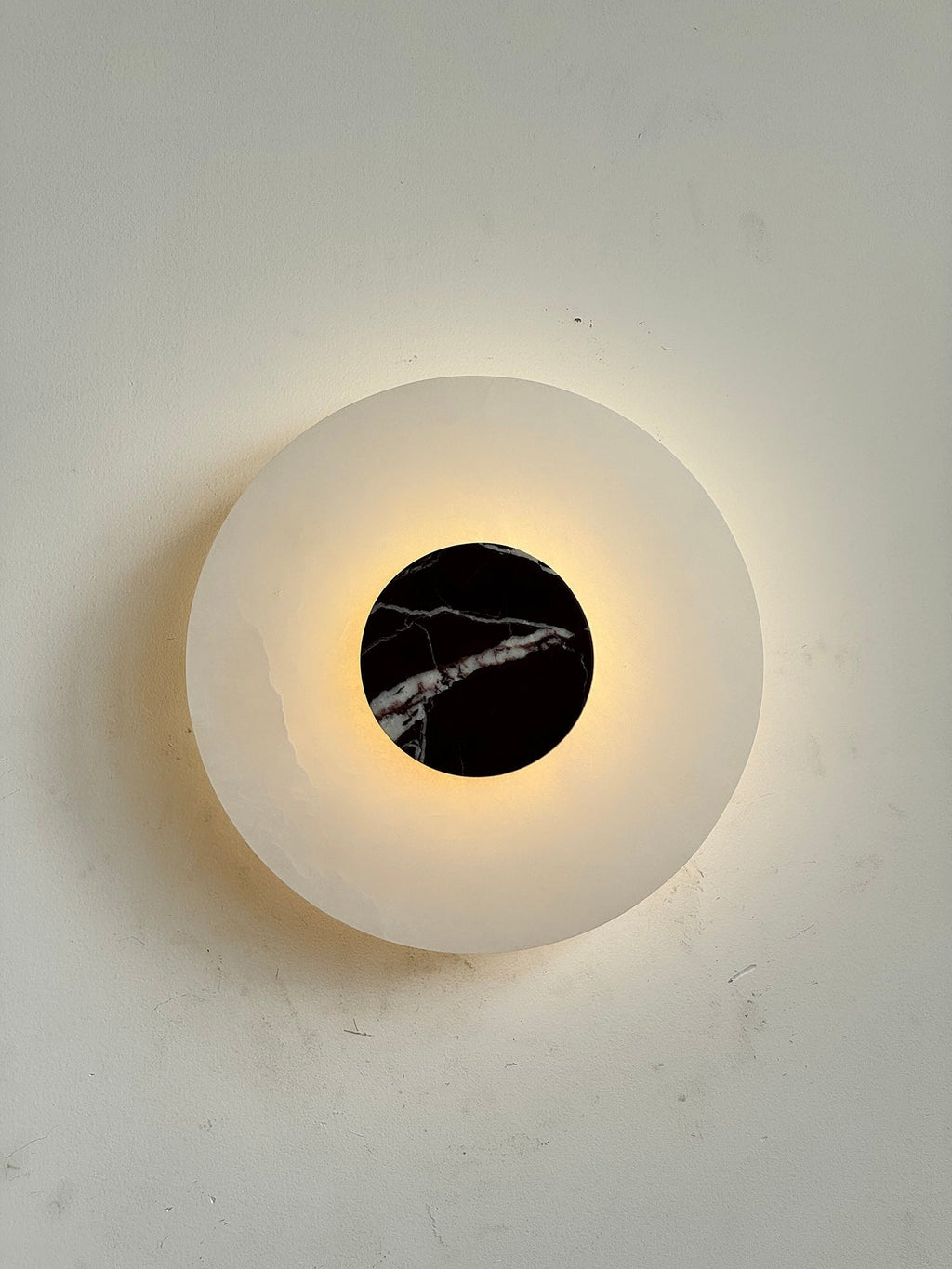 The Clérachic wall lamp lit head-on, warm light ringing the black marble disc and pooling across the alabaster plate.