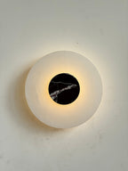 The Clérachic wall lamp lit head-on, warm light ringing the black marble disc and pooling across the alabaster plate.
