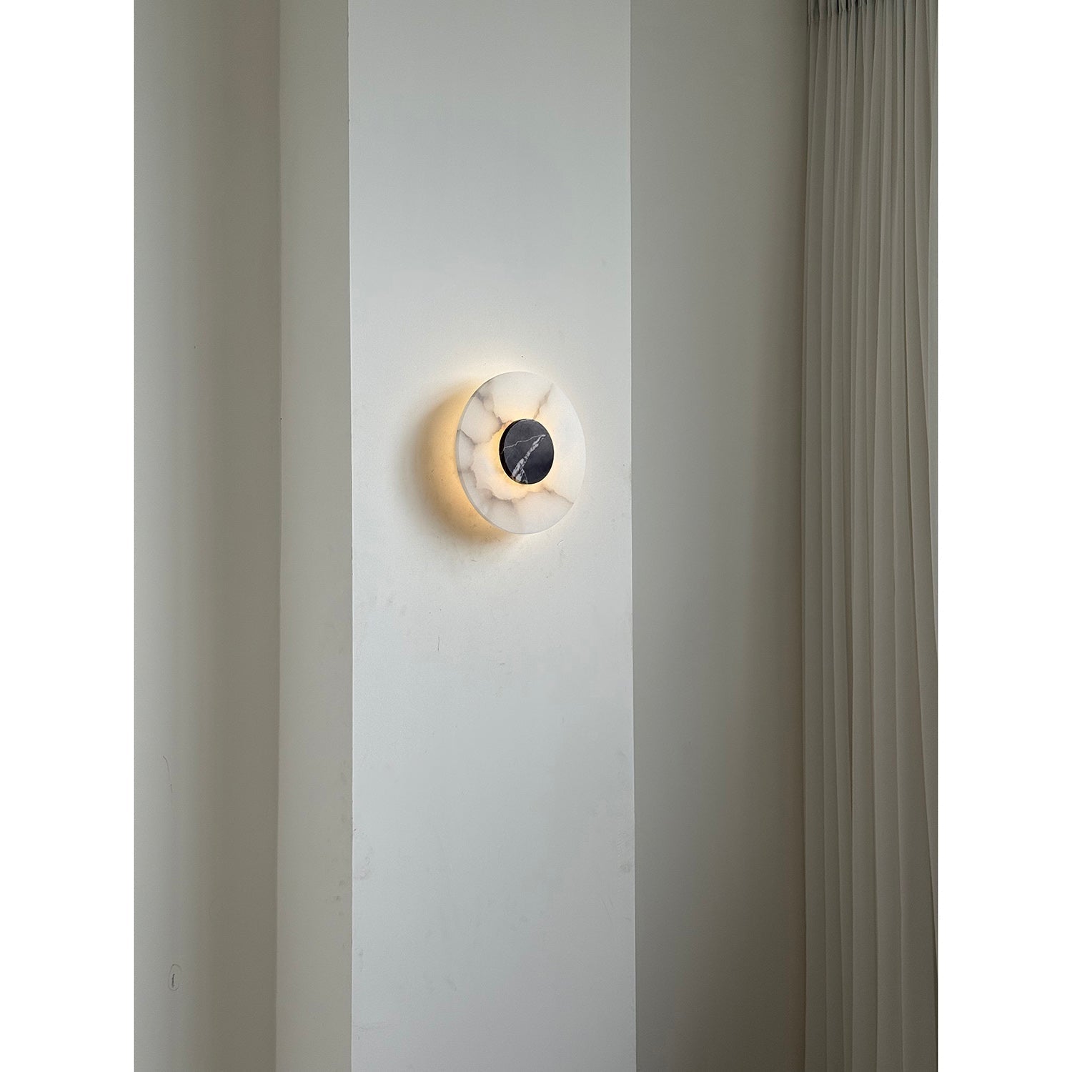 The Clérachic wall lamp off against a pale pier, its veined alabaster plate catching daylight beside a pleated curtain.