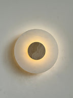 Clérachic wall lamp lit head-on with a beige travertine disc at its centre, warm halo spreading over the plaster.