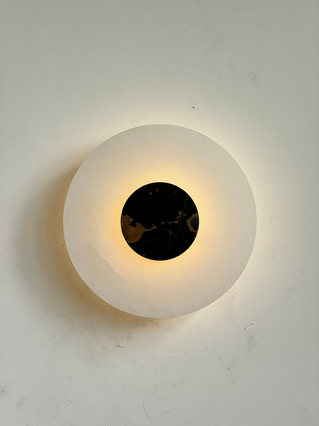 The Clérachic wall lamp lit square-on, warm light escaping around a black and gold marble disc.