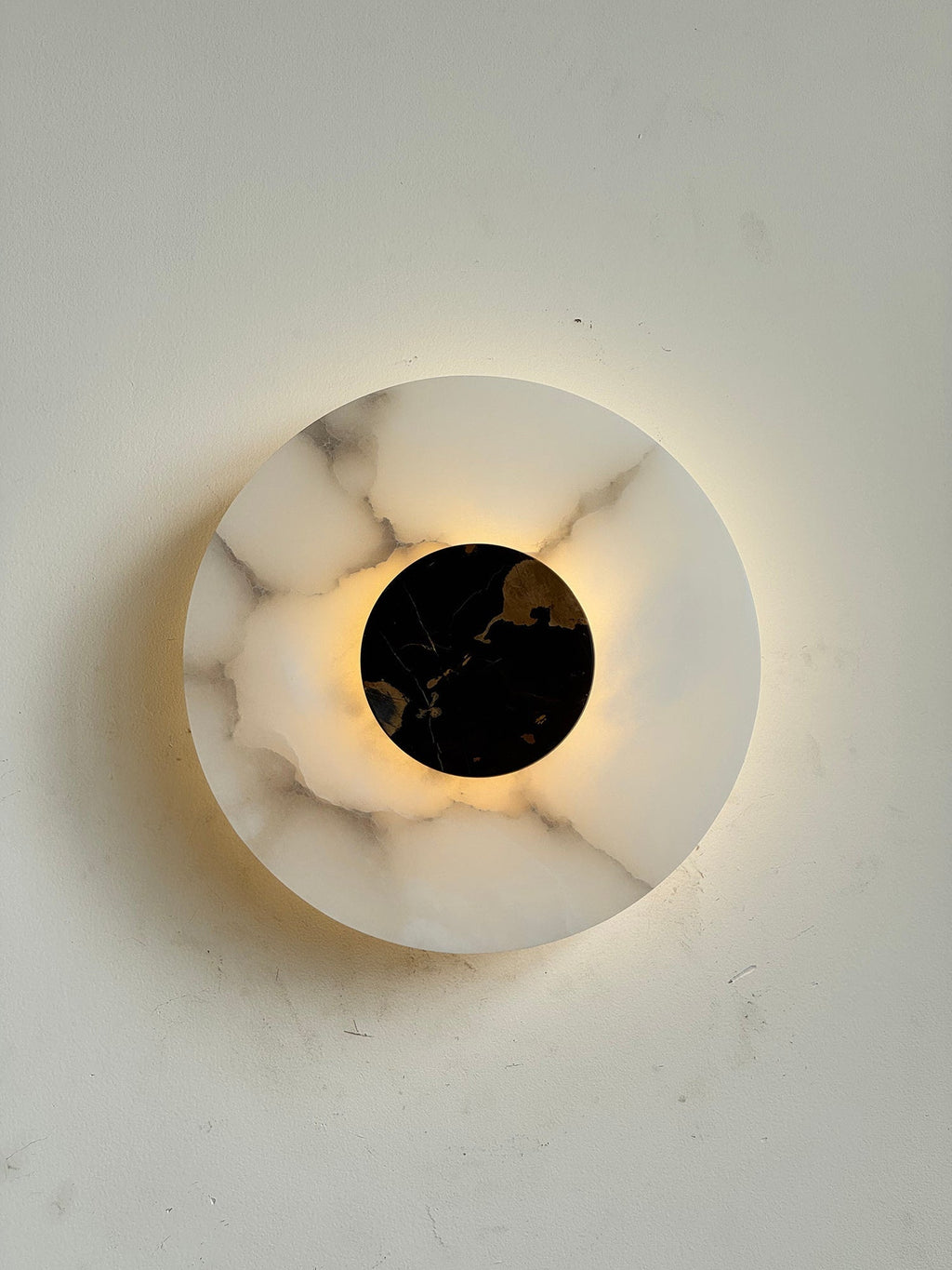 Clérachic wall lamp lit head-on, a bright corona around the black marble disc picking out amber veining in the stone.