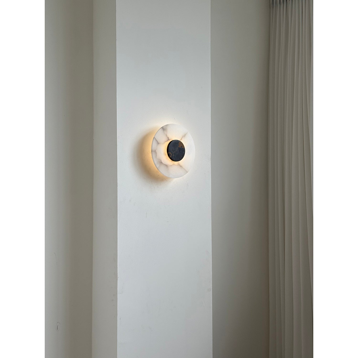 Clérachic wall lamp unlit on an interior pier, the veined alabaster plate framed by a pleated curtain to one side.