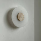 Clérachic wall lamp unlit at an angle, its beige travertine disc set off-centre on the smooth alabaster plate.