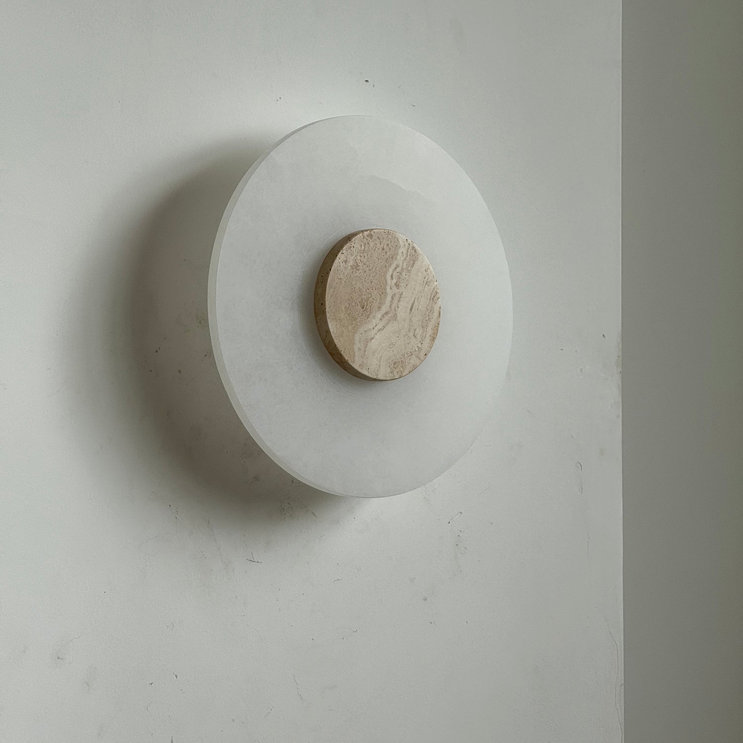 Clérachic wall lamp unlit at an angle, its beige travertine disc set off-centre on the smooth alabaster plate.