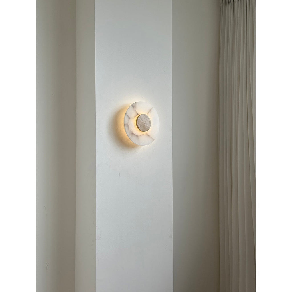 Clérachic wall lamp lit on a pale pier, the travertine centre held inside a soft ring of warm light.