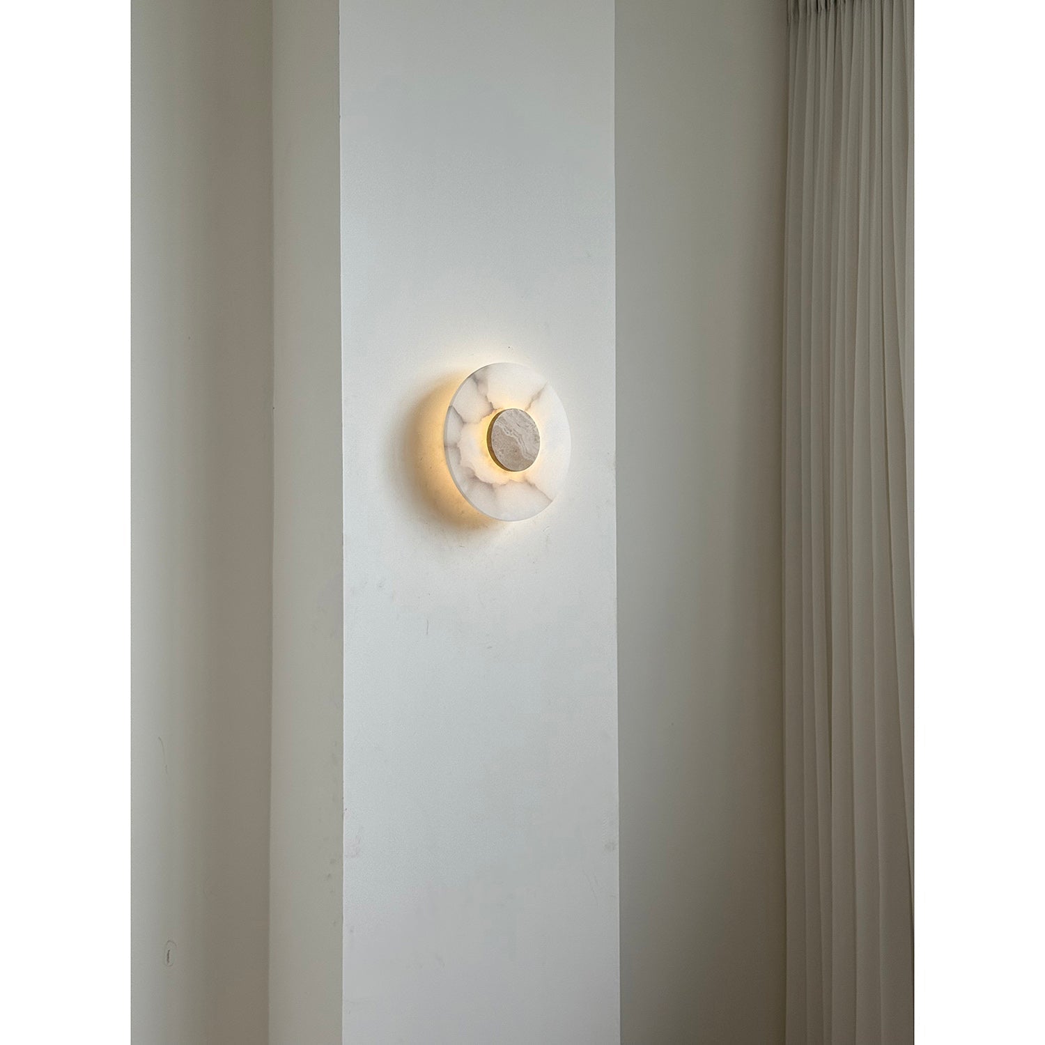 Clérachic wall lamp lit on a pale pier, the travertine centre held inside a soft ring of warm light.