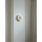 The Clérachic wall lamp unlit in a room, its travertine disc a small warm dot against white alabaster and plaster.
