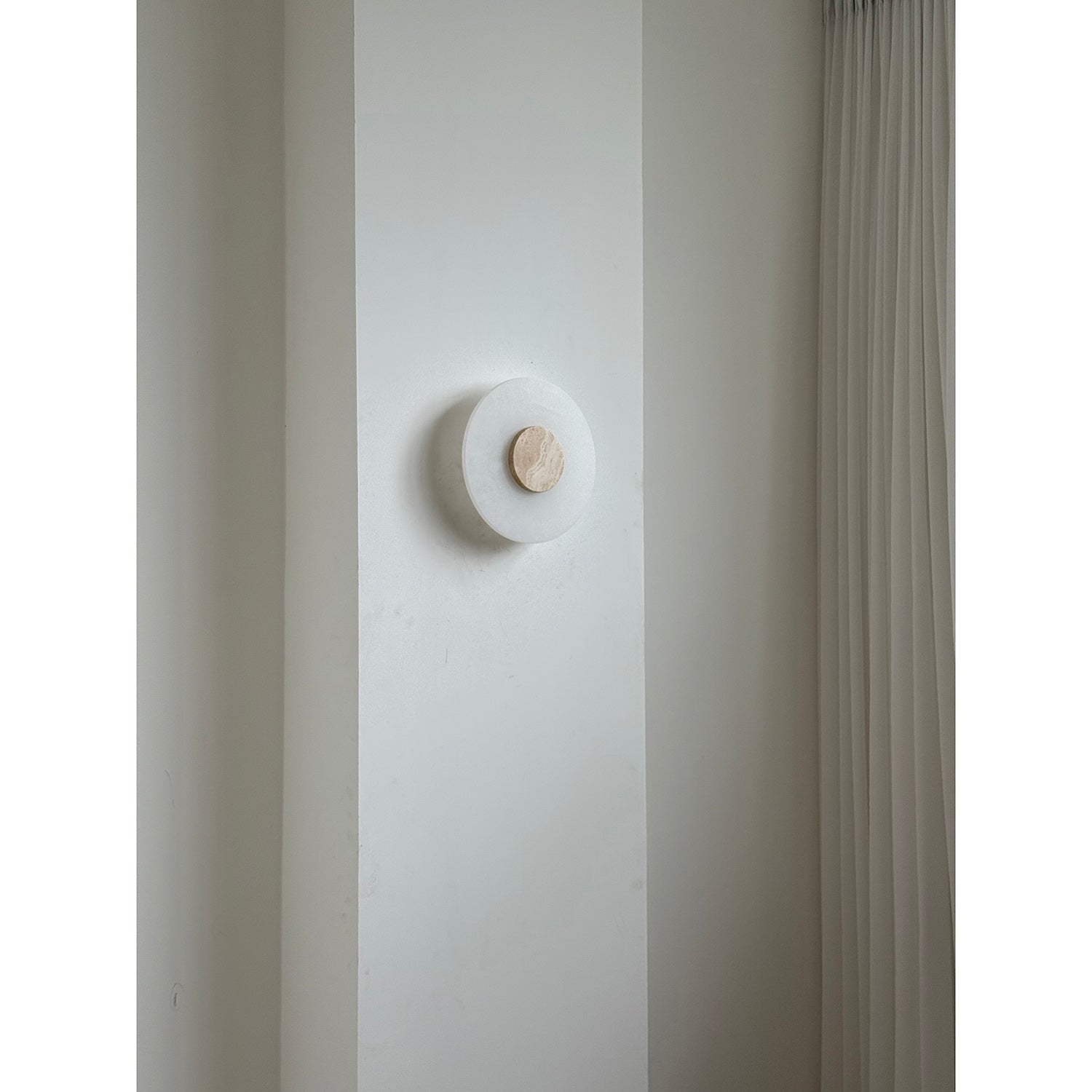 The Clérachic wall lamp unlit in a room, its travertine disc a small warm dot against white alabaster and plaster.