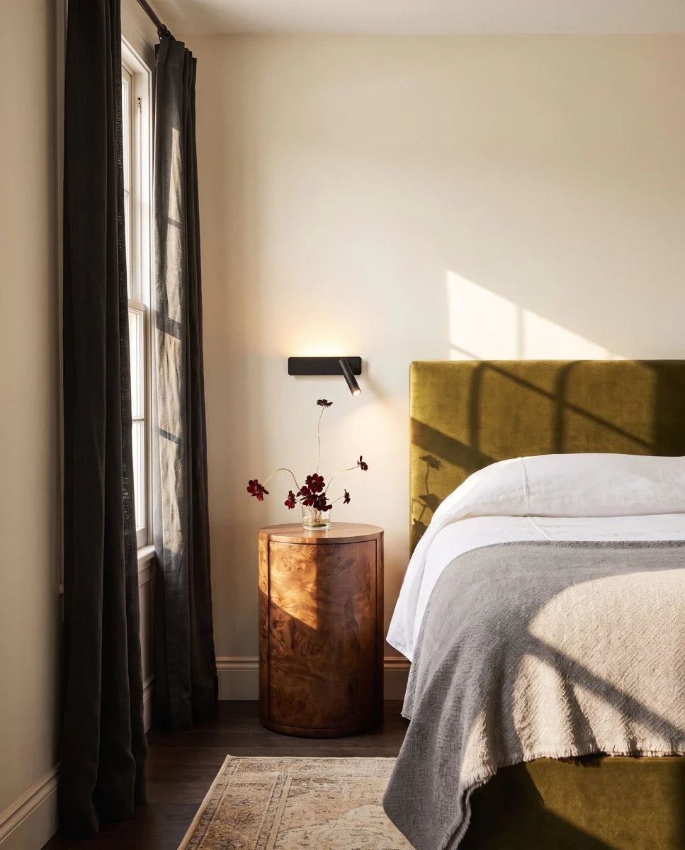 Clérachic black reading wall light above a burr-wood bedside table, beside an olive velvet headboard in a sunlit bedroom.