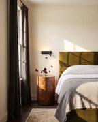 Clérachic black reading wall light above a burr-wood bedside table, beside an olive velvet headboard in a sunlit bedroom.