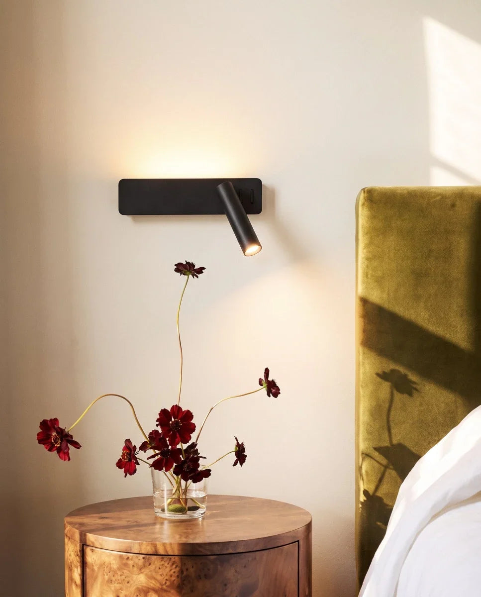 Close view of the Clérachic black wall reading light glowing above a glass vase of dark red flowers and a round wooden table.
