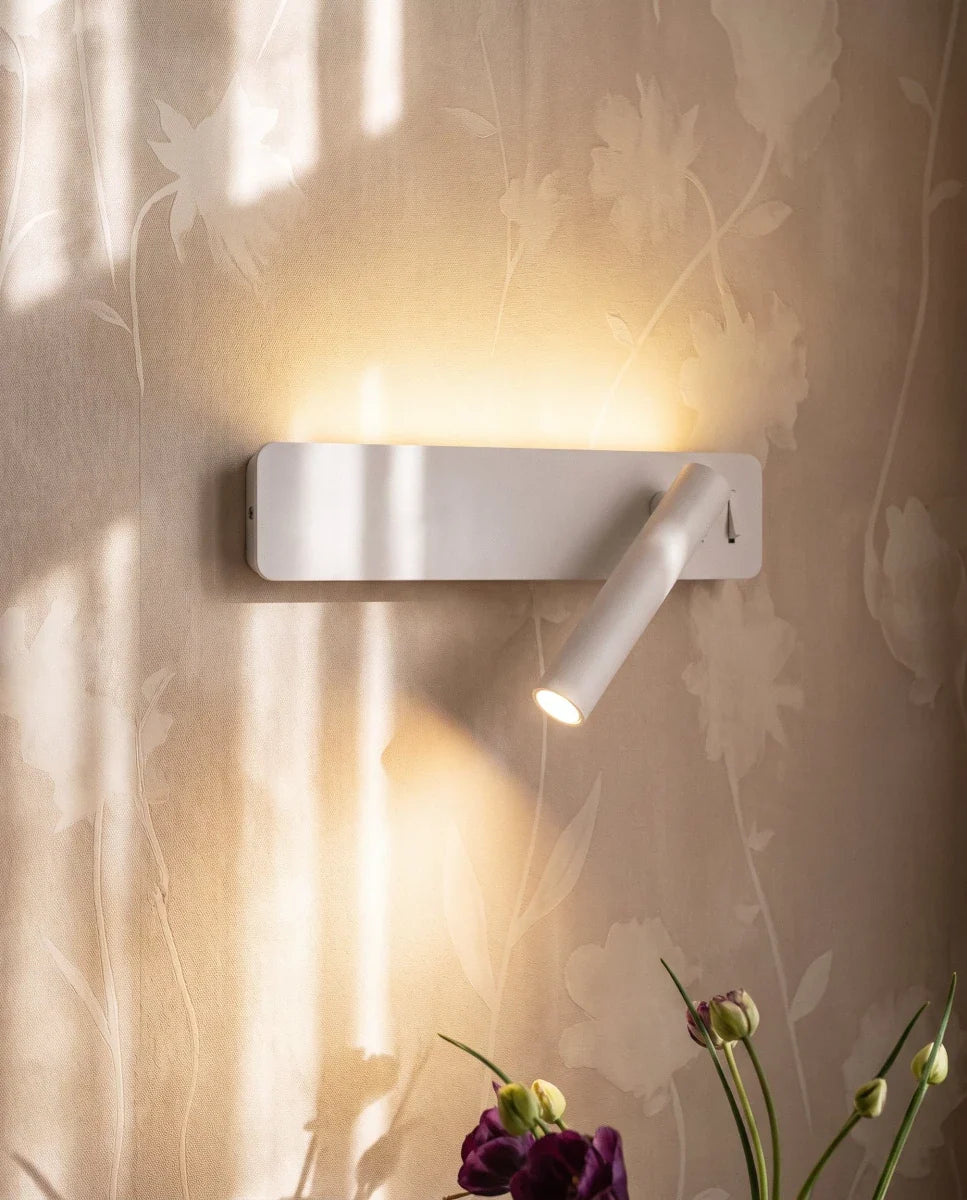 Clérachic white wall reading light glowing against floral wallpaper, its cylindrical arm angled down with a small switch at the side.