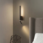 Clérachic slim black wall light with a warm vertical glow and a downward reading spot beside a grey studded headboard.