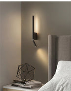 Clérachic slim black wall light above a bedside table with books and a wire sculpture, beside a grey studded headboard.