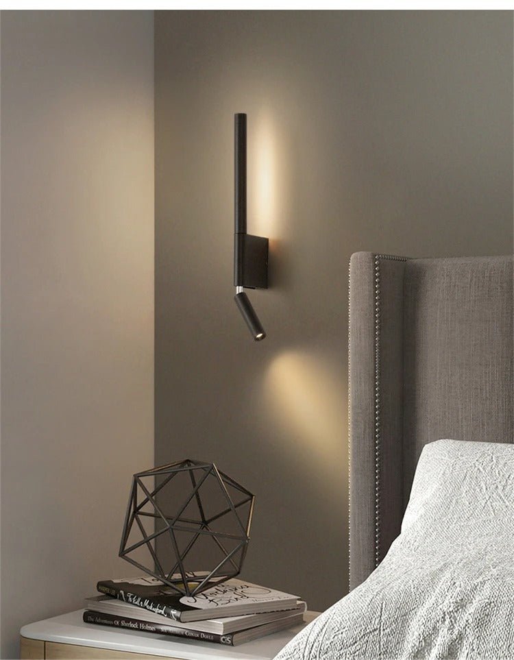 Clérachic slim black wall light above a bedside table with books and a wire sculpture, beside a grey studded headboard.