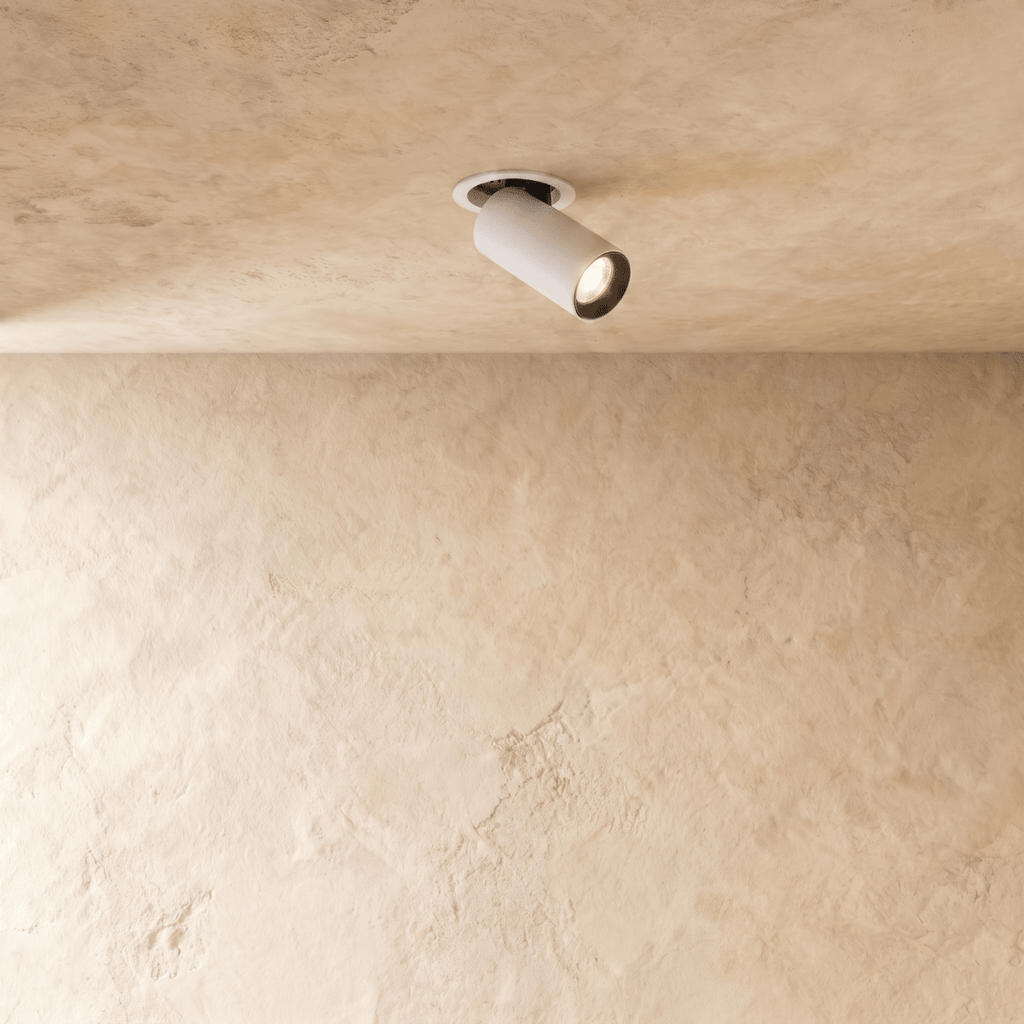 Clérachic recessed spotlight in white, its cylindrical head angled out of a round trim on a limewashed plaster ceiling, lit.