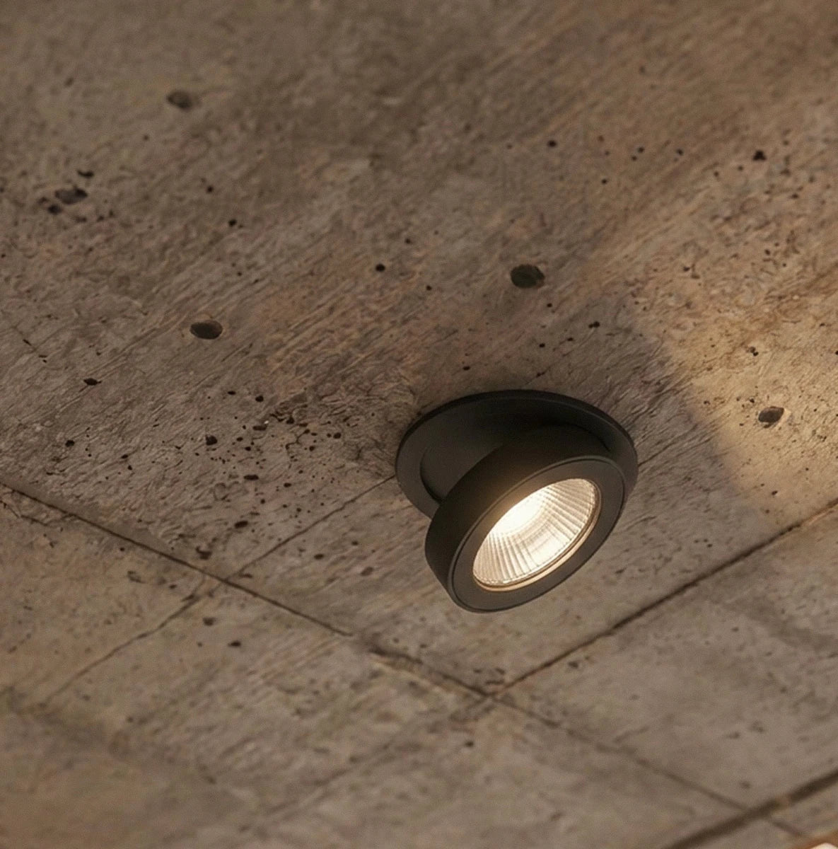 Clérachic recessed downlight in black, lit close-up on a raw concrete ceiling showing the ridged optic face inside a smooth bezel.