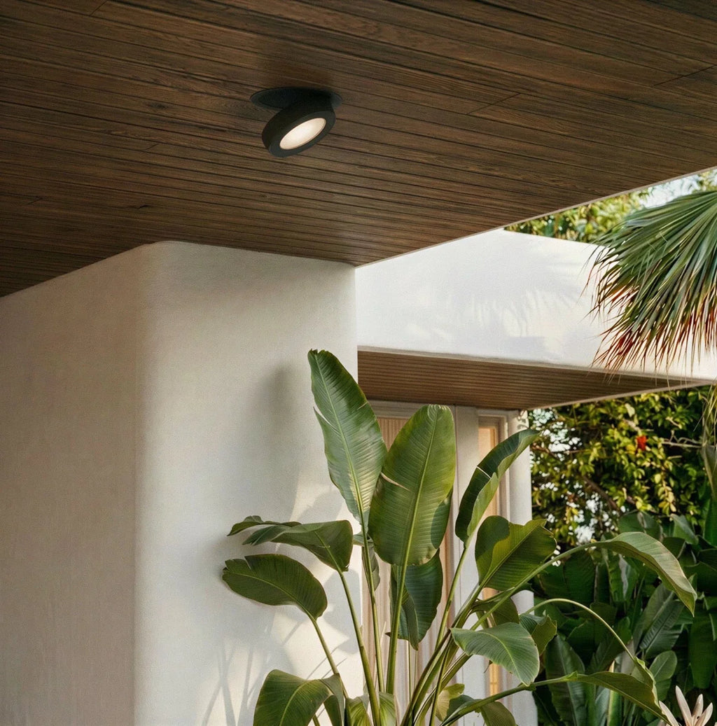 Clérachic recessed downlight in black with a frosted lens, lit in a wood-slat ceiling beside a white column and a banana-leaf plant.