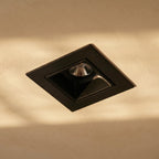 Close view of a Clérachic square black recessed downlight with a deep reflector in a plaster ceiling washed by warm light.