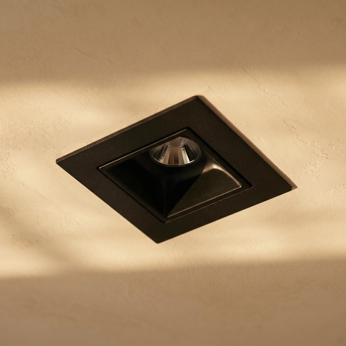 Close view of a Clérachic square black recessed downlight with a deep reflector in a plaster ceiling washed by warm light.
