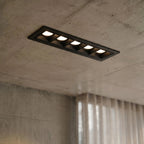 Clérachic five-head black linear recessed downlight, lit white, set into a raw concrete ceiling beside sheer curtains.