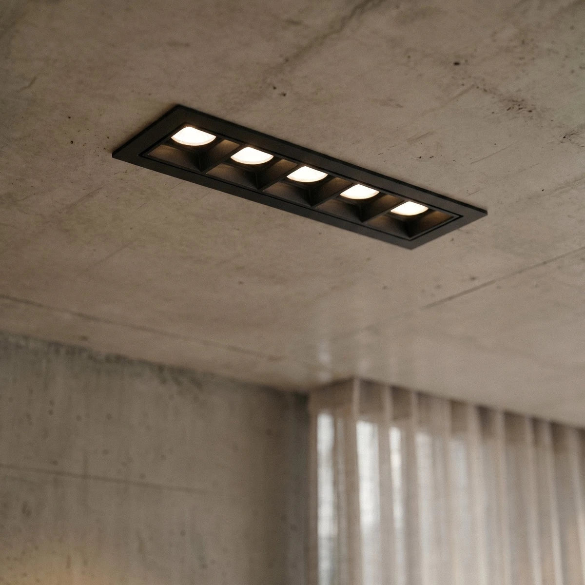 Clérachic five-head black linear recessed downlight, lit white, set into a raw concrete ceiling beside sheer curtains.