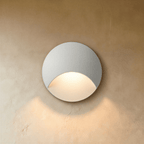 Clérachic recessed step light in white with a crescent-cut face, warm glow washing softly down a plaster stairwell wall.