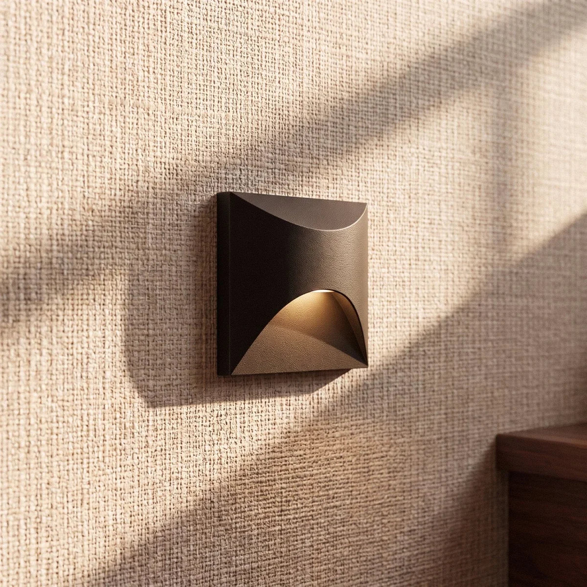 Clérachic recessed step light in matte black with a square face, warm glow lit close against a woven wall covering.