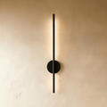 Clérachic slim black vertical wall lamp on a round backplate, glowing warm along its length on a limewash wall.