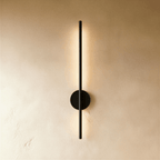 Clérachic slim black vertical wall lamp on a round backplate, glowing warm along its length on a limewash wall.