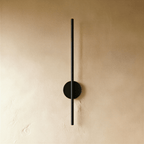 Clérachic slim black vertical wall lamp with a round backplate, switched off against a warm textured plaster wall.