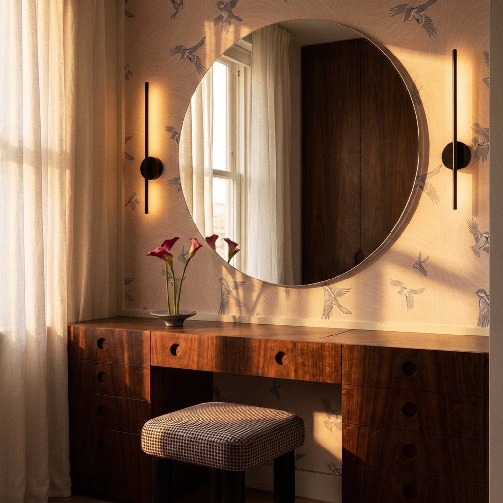 Pair of Clérachic slim black wall lamps flanking a large round mirror above a walnut dressing table and houndstooth stool.