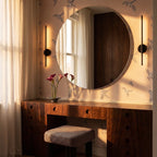 Pair of Clérachic slim black wall lamps flanking a large round mirror above a walnut dressing table and houndstooth stool.