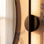 Close-up of the Clérachic black wall lamp, its round backplate and slim rod beside a mirror edge and bird wallpaper.