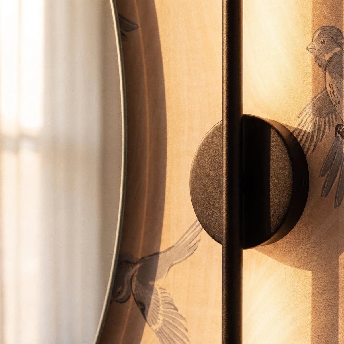 Close-up of the Clérachic black wall lamp, its round backplate and slim rod beside a mirror edge and bird wallpaper.