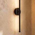 Close-up of the Clérachic black wall lamp, its round backplate and rod end with warm light spreading on plaster.