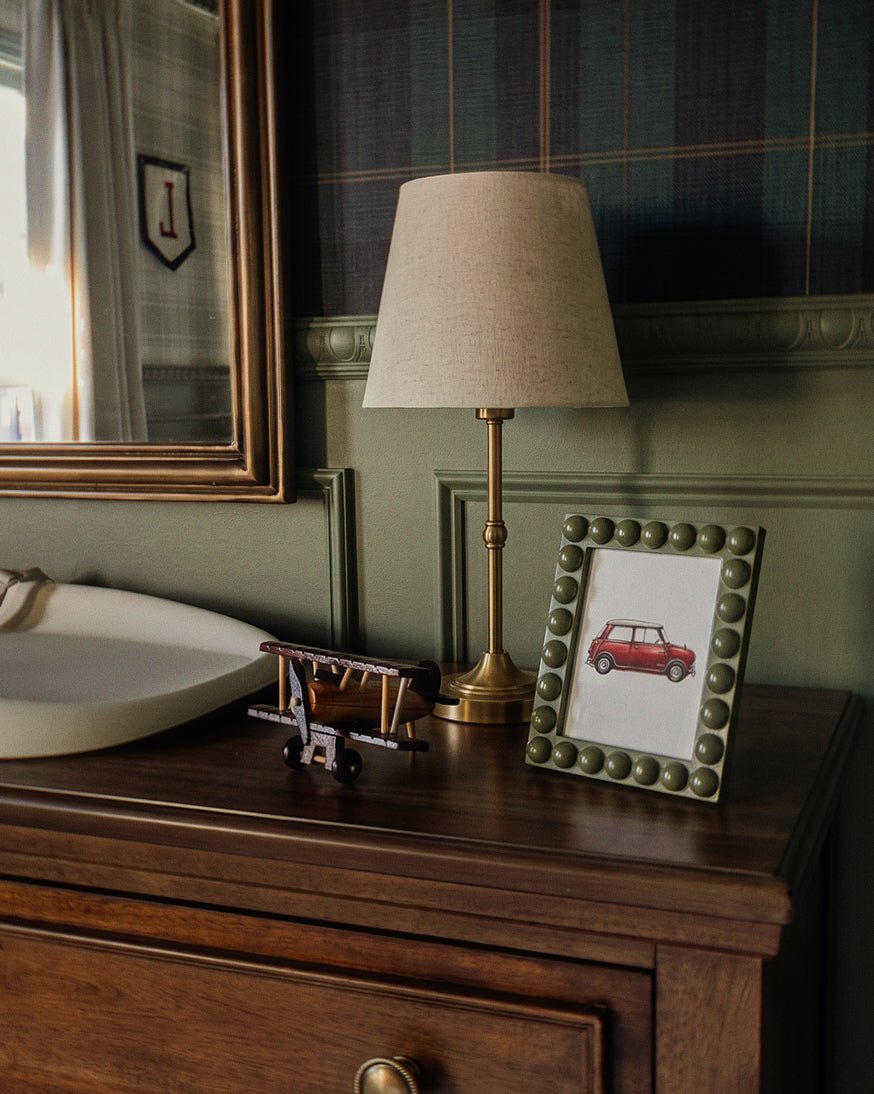 Clérachic brushed gold table lamp with a tapered linen shade on a dark wood dresser beside a framed print and a toy plane.