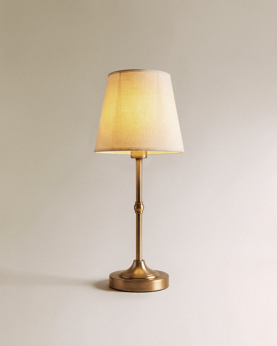 Clérachic table lamp with a slim brushed gold stem, round weighted base and tapered white fabric shade, glowing warm.