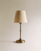 Clérachic table lamp with a brushed gold stem and weighted round base, topped by a tapered natural linen shade, switched off.