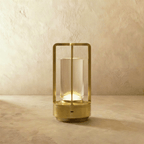 Clérachic brass table lamp: an open metal frame around a clear glass tube, glowing warm on a limewash-textured backdrop.