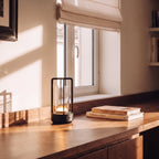 Clérachic black table lamp glowing on a walnut desk beside two books, in low window sunlight with a roman blind behind.