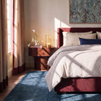 Clérachic brass table lamp lit on a burl-wood bedside table next to a vase of white calla lilies, with a burgundy bed alongside.