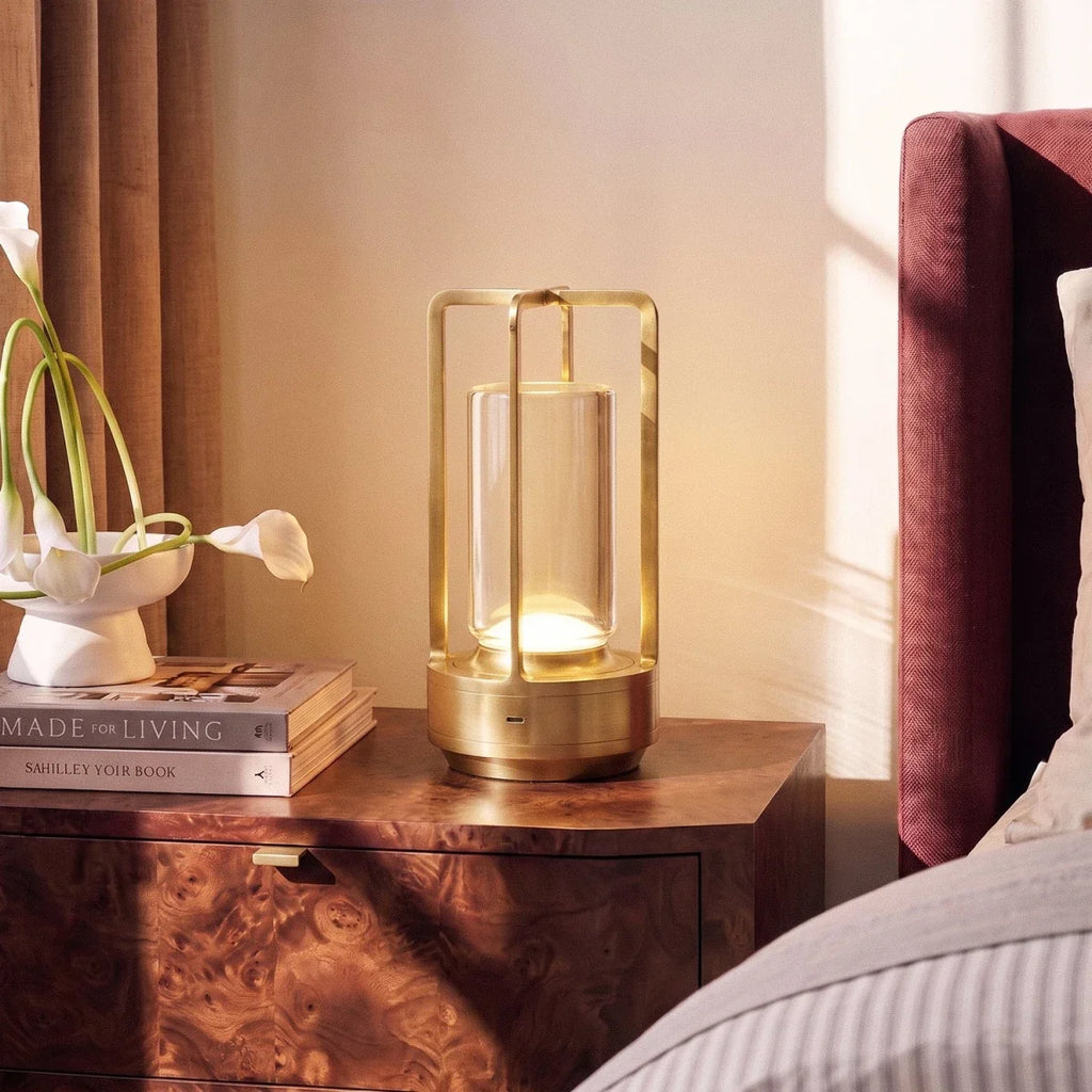 Clérachic brass table lamp glowing on a burl-wood bedside table, with calla lilies in a white dish and a stack of books beside it.