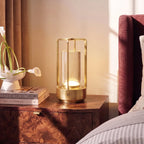 Clérachic brass table lamp glowing on a burl-wood bedside table, with calla lilies in a white dish and a stack of books beside it.