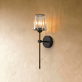 Clérachic black wall sconce on a slim rod with a fluted clear glass shade, glowing warm against a limewash wall.