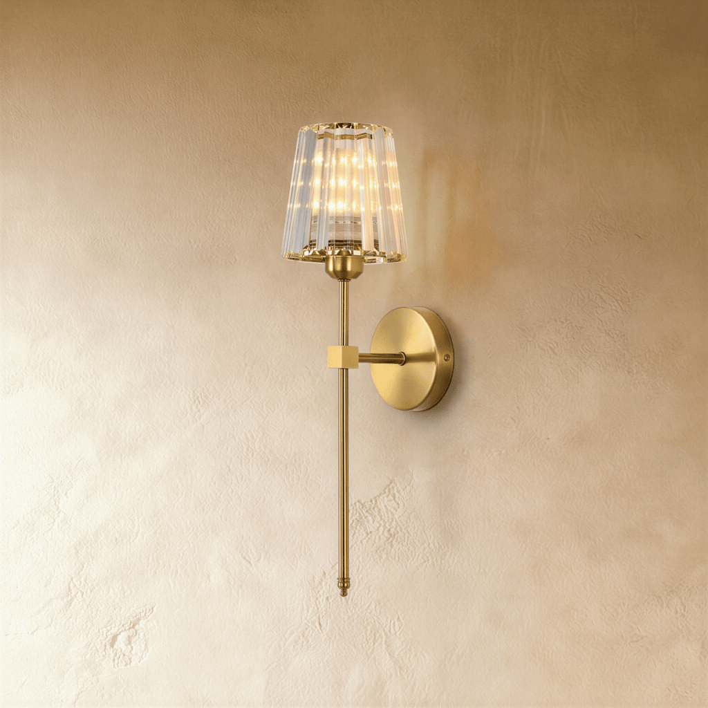 Clérachic brass-gold wall sconce with a faceted clear glass shade on a slim rod and round backplate, lit on a pale wall.