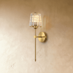Clérachic brass-gold wall sconce with a faceted clear glass shade on a slim rod and round backplate, lit on a pale wall.